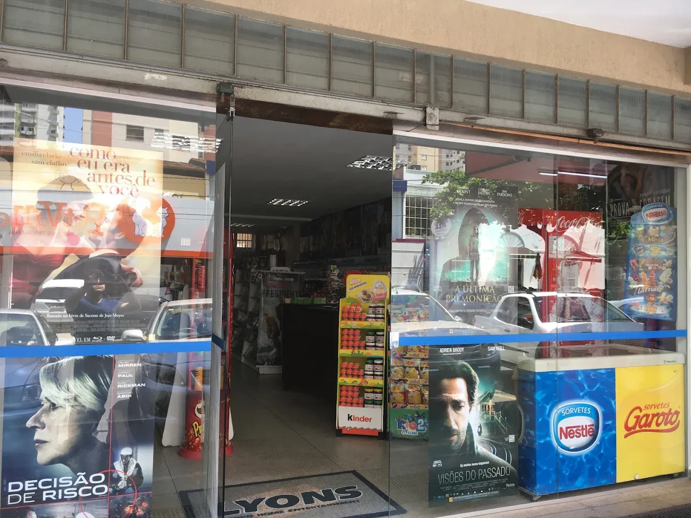 Fachada da Lyons Store na época da videolocadora, com cartazes de filmes na vitrine e o tapete com o nome Lyons na entrada